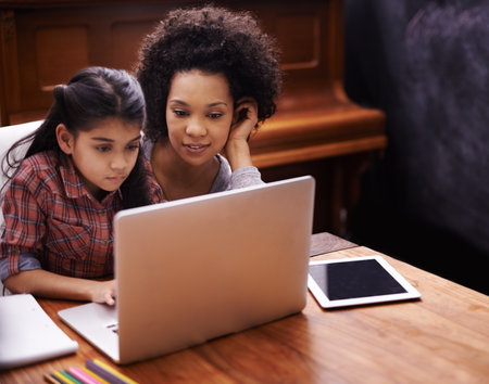 Allowing her to do it on her own. Shot of a little girl using a laptop to do her homework while her mother sits by.の写真素材
