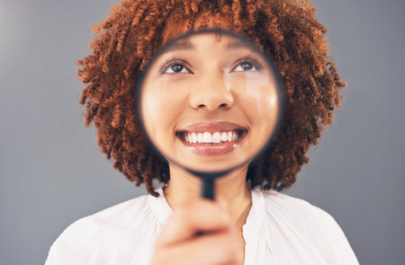 Funny, smile and black woman with a magnifying glass on face isolated on a grey studio background. Happy, comic and facial zoom of a girl with a magnifier for comedy, happiness and discoveryの写真素材