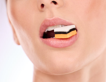 Liquorice, woman eating and mouth with makeup and cosmetics in an isolated studio. White background, sweet food and candy product with hungry female model with cheat meal and sugar for dessertの写真素材