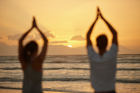Appreciating the beauty of nature while doing yoga. Rearview shot of a couple doing yoga on the beach at sunset.の写真素材