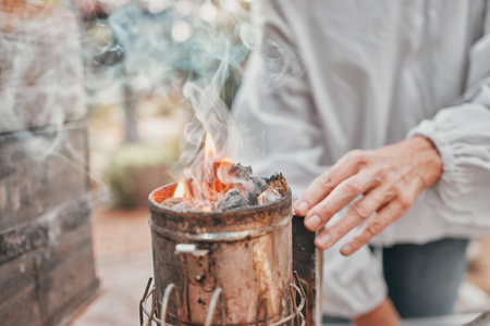 Agriculture, fire and hands of a beekeeper in production of honey with smoke in container....