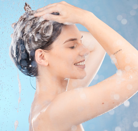 Woman, shampoo and shower for hair care, clean or hygiene on blue studio background. Young female, girl or lady wet, water or bubbles for natural beauty, morning routine or skincare to relax or smileの写真素材