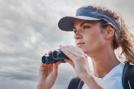 Woman, binoculars and hiking in nature, travel or outdoors vacation, holiday or trip in Canada. Freedom, cloudy sky and female on adventure, sightseeing or trekking and looking with field glasses.の写真素材