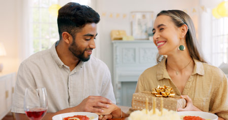 Interracial couple, gift and celebrate birthday being happy, kiss and smile in home at table with cake. Love, man and woman being content, romantic and present being cheerful celebration together.の写真素材