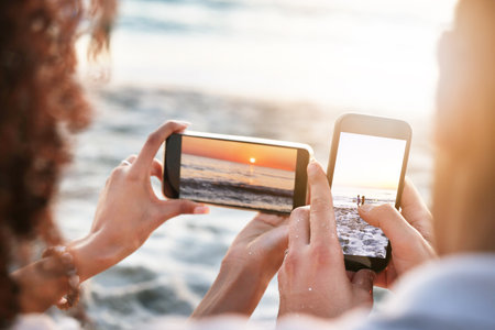 Phones, beach and couple taking a picture of the sunset while on summer vacation or weekend trip. Technology, adventure and hands of man and woman with cellphones taking photo by the ocean on holidayの写真素材