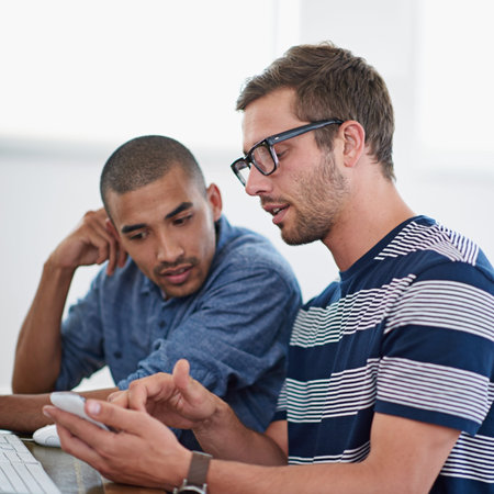 Going through the project to do list. two coworkers looking at a smartphone while sitting at a desk...
