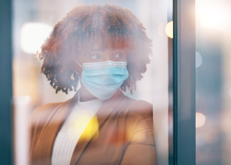 Covid, window and thinking with a business black woman in her office, standing by a glass pane alone. Idea, safety and mask with a female employee in her workplace during the corona virus pandemicの写真素材