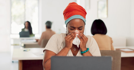 Black woman, sneeze and tissue blowing nose for sick, ill or flu by laptop at the office desk. African American woman employee with cold symptoms, virus or illness by computer at the workplaceの写真素材