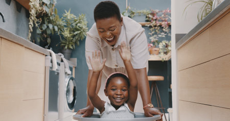 Playful black mother pushing her daughter around in a laundry basket at home. Young woman and her child playing and having fun while spending time at homeの写真素材