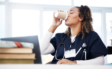 Medical student, technology and drinking coffee in hospital studying burnout, education books research or wellness learning. Tired, nurse or healthcare woman with drink, laptop or medicine internshipの写真素材