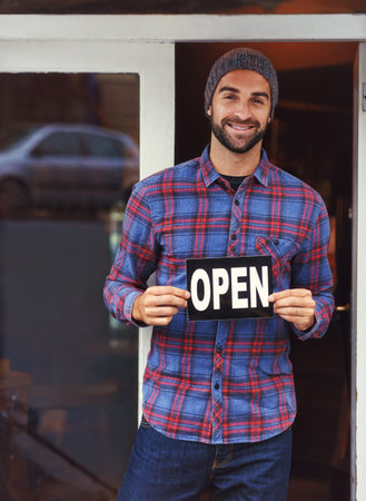 Come have a look inside. a handsome young man standing next to his business and holding up the open...