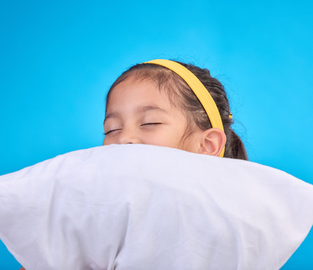 Little girl, pillow and sleep in a studio feeling tired, fatigue and ready for dreaming. Isolated, blue background and happy young child with pillows and closed eyes for sleeping, rest and napの写真素材