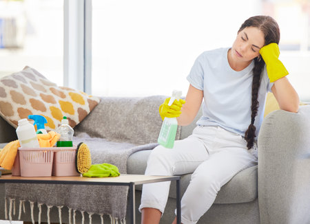 Tired, sad and spring cleaning woman in living room with burnout, stress and fatigue from housekeeping, maintenance and routine tasks. Exhausted, sleepy and female cleaner, maid and headache problemの写真素材