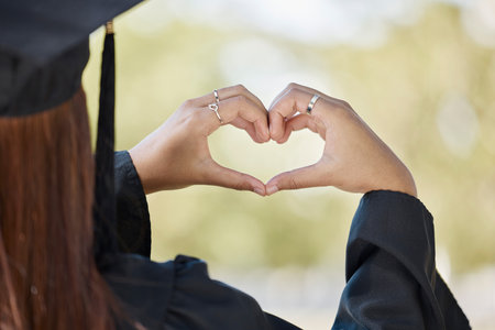 Student graduation, heart hand sign and education achievement outdoor with blurred background. University, college success and love emoji hands gesture from education event and graduate womanの写真素材