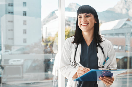 Woman, doctor and smile writing on clipboard by window for healthcare planning, strategy or notes. Happy female medical expert smiling with paperwork, prescription or medicare details at hospitalの写真素材