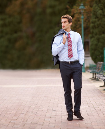 Taking an after-work stroll in the park. A handsome businessman walking outside with his jacket over his shoulder.の写真素材