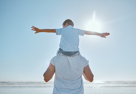 Family, beach and summer vacation for freedom with a child on shoulder of father with hands outstretched for happiness against blue sky. Man and kid son together at sea for trust, nature and peaceの写真素材