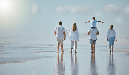 Family, beach and men with women and child from back with blue sky on outdoor summer vacation. Happy family mother, father and kid with piggy back fun walking in sun and quality time together at sea.の写真素材