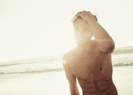 Catching the last rays. A good-looking man standing with his hand in his hair on the beach at sunset.の写真素材