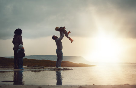 Sunset, view and a playing family on the beach together on a beautiful summer evening outdoor. Earth, water or nature with a mother, father and children bonding by the ocean or sea at the coastの写真素材