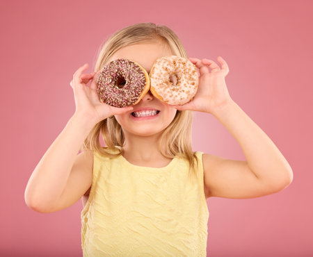 Child, donut over eyes and smile in studio with sweet snack in hands on a pink background. Girl kid model with happiness, creativity and comic face holding food in hand isolated on color and spaceの写真素材
