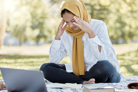 Stress, Islamic and woman student with a headache while studying in a park feeling frustrated and sad outdoors. Laptop, sick and muslim female with a hijab annoyed while learning or readingの写真素材
