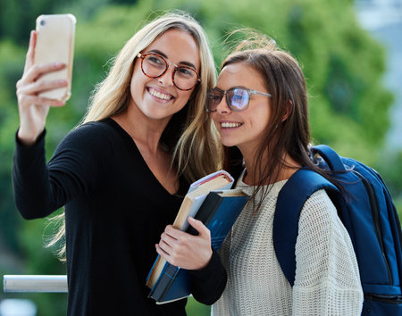 Selfie, women and students on campus, goals and happiness with success, cheerful and books. Females, girls and academics with smartphone, social media and connection outdoor and picture for memoriesの写真素材