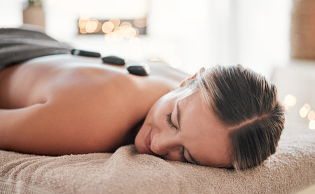 Woman, stone and back on bed in spa treatment for relaxation, stress relief or massage at resort. Happy female relaxing with hot rocks in physical therapy for body healing, zen wellness or skincareの写真素材