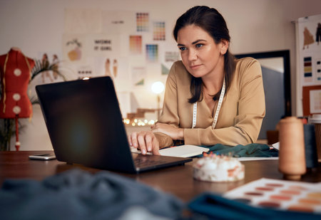 Laptop, fashion and design with a woman tailor or seamstress typing a proposal in her workshop for...