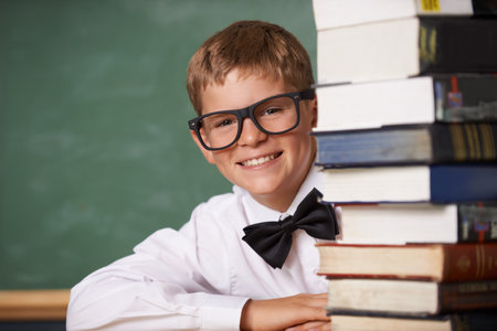 This is any bookworms dream. A young boy wearing glasses and a bow-tie smiling at the camera from behind a stack of books.の写真素材