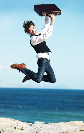 The freedom of being self-employed. Happy young businessman jumping up holding his briefcase in the air with the ocean in the background.の写真素材