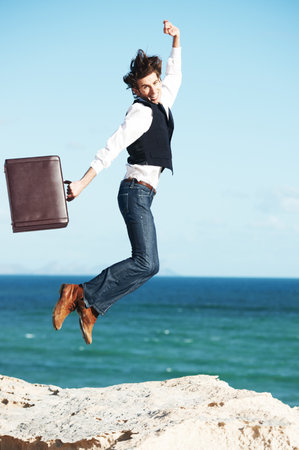 Its great to be out of the office. Young man holding a briefcase and jumping in the air with the ocean in the background.の写真素材