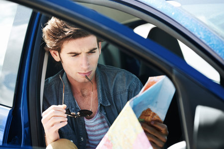 Navigating my journey. Young man sitting in his car and inspecting a map while holding his...