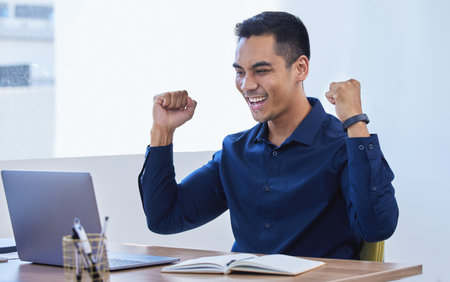 Success is inevitable if you keep at it. a young businessman cheering while working on a laptop in an office.の写真素材