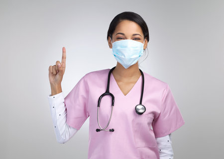 Things are looking up. Cropped portrait of an attractive young female healthcare worker wearing a mask and pointing towards copyspace in studio against a grey background.の写真素材