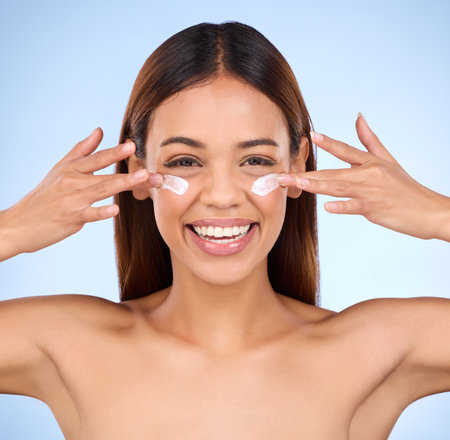 Skincare, beauty and portrait of happy woman with eye cream, anti aging or skin care on blue background. Cosmetics, facial lotion and face of hispanic model with smile and collagen product in studio.の写真素材