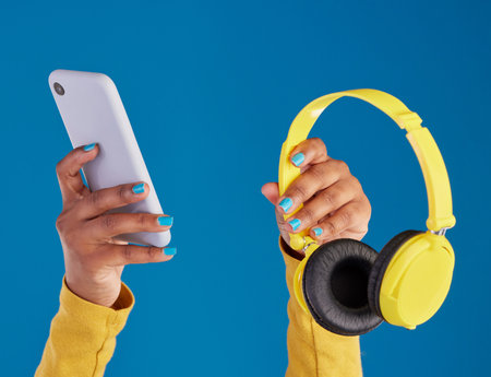Closeup, hands and smartphone with headphones, streaming music and audio against blue studio background. Zoom, hand and technology with cellphone, headset or radio with sounds, internet or connectionの写真素材