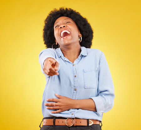 Pointing, laughing and fun with a black woman in studio on a yellow background enjoying a funny joke. Comic, comedy and laughter with an attractive young female feeling silly, goofy or playfulの写真素材