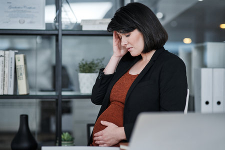 Why am I getting headaches this late into my pregnancy. an attractive businesswoman sitting and feeling stressed while holding her pregnant tummy in the office.の写真素材