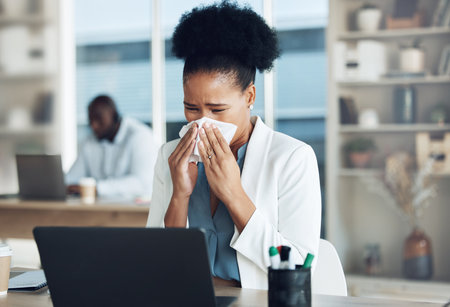 Work, healthcare and black woman at desk blowing nose with tissue paper from flu, cold or hay fever. Sick, exhausted office employee with allergy and sinus problem, health risk from illness at laptopの写真素材