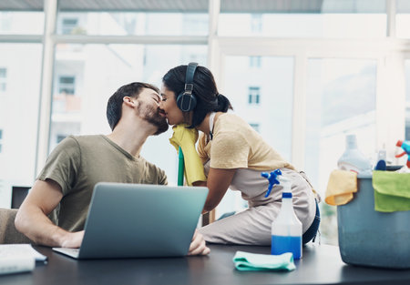 For anyone who wants to know why freelancing is awesome. a young woman kissing her husband while she cleans the house and he uses a laptop.の写真素材