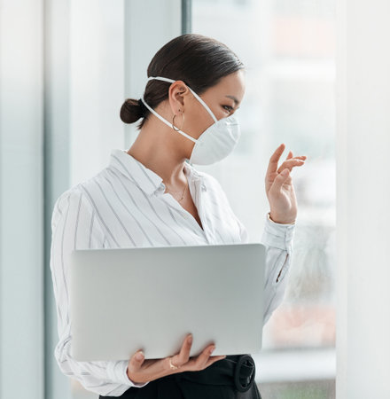 If we cant connect in person lets connect online. a masked young businesswoman holding a laptop and waving out of a window in a modern office.の写真素材