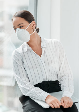 The landscape of business sure has changed. a masked young businesswoman looking out of a window in a modern office.の写真素材