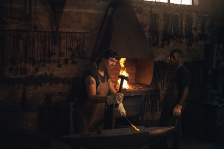 Strike while the iron is hot. a young woman hitting a hot metal rod with a hammer in a foundry.