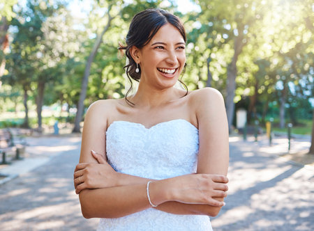 Beautiful mixed race bride wearing a white strapless dress while standing with her arms crossed in nature on a sunny day. Carefree bride smiling and dreaming about her her future as a wifeの写真素材