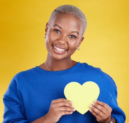 Paper heart, portrait and happy black woman in studio, background and color backdrop. Smile, female model and yellow shape for love, wellness and caring support of peace, romance and sign of kindnessの写真素材