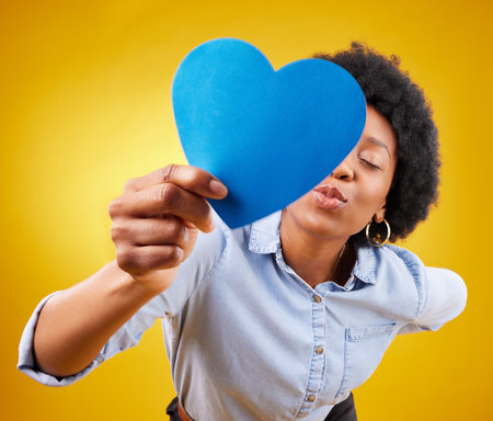 Paper, heart and kissing with black woman in studio for romance, positive and emotion. Happiness, love shape and giving with female isolated on yellow background for date, feelings and affectionateの写真素材