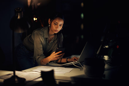 I had that message saved on my phone somewhere...a young businesswoman using a cellphone while working on a laptop in an office at night.の写真素材