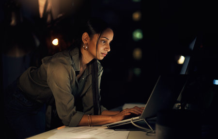 Keeping her mind on the deadline. a young businesswoman working on a laptop in an office at night.の写真素材