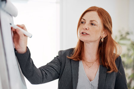 Giving direction and instruction for her team to follow. a mature businesswoman using a whiteboard while giving a presentation in an office.の写真素材